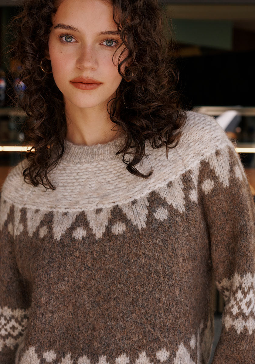 A young person with curly brown hair and blue eyes wears a Princess Goes Hollywood Jacquard pullover with raglan sleeves in a wool-alpaca mix and Nordic design, gazing neutrally at the camera against a softly blurred indoor background.
