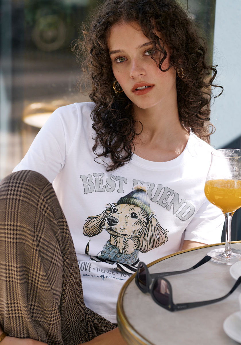 A young woman with curly brown hair sits outdoors, wearing a Princess Goes Hollywood cotton T-shirt with a cute dachshund print and “BEST FRIEND” text. She pairs it with plaid pants and gold hoops, glancing at the camera. An orange drink and sunglasses are on the table.