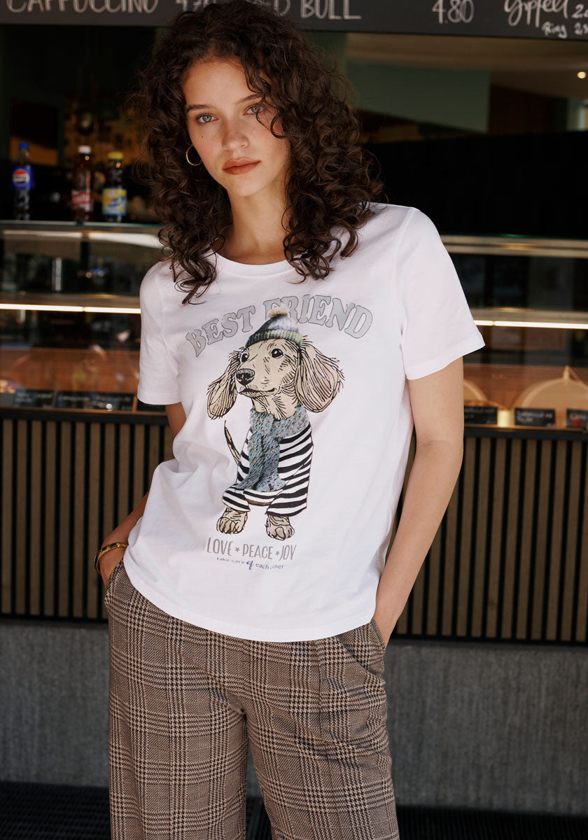 A woman with curly brown hair stands at a café counter in plaid pants and a Princess Goes Hollywood cotton T-shirt featuring a dachshund print, a maritime vibe, and the words BEST FRIEND LOVE PEACE JOY. Her hands are in her pockets.