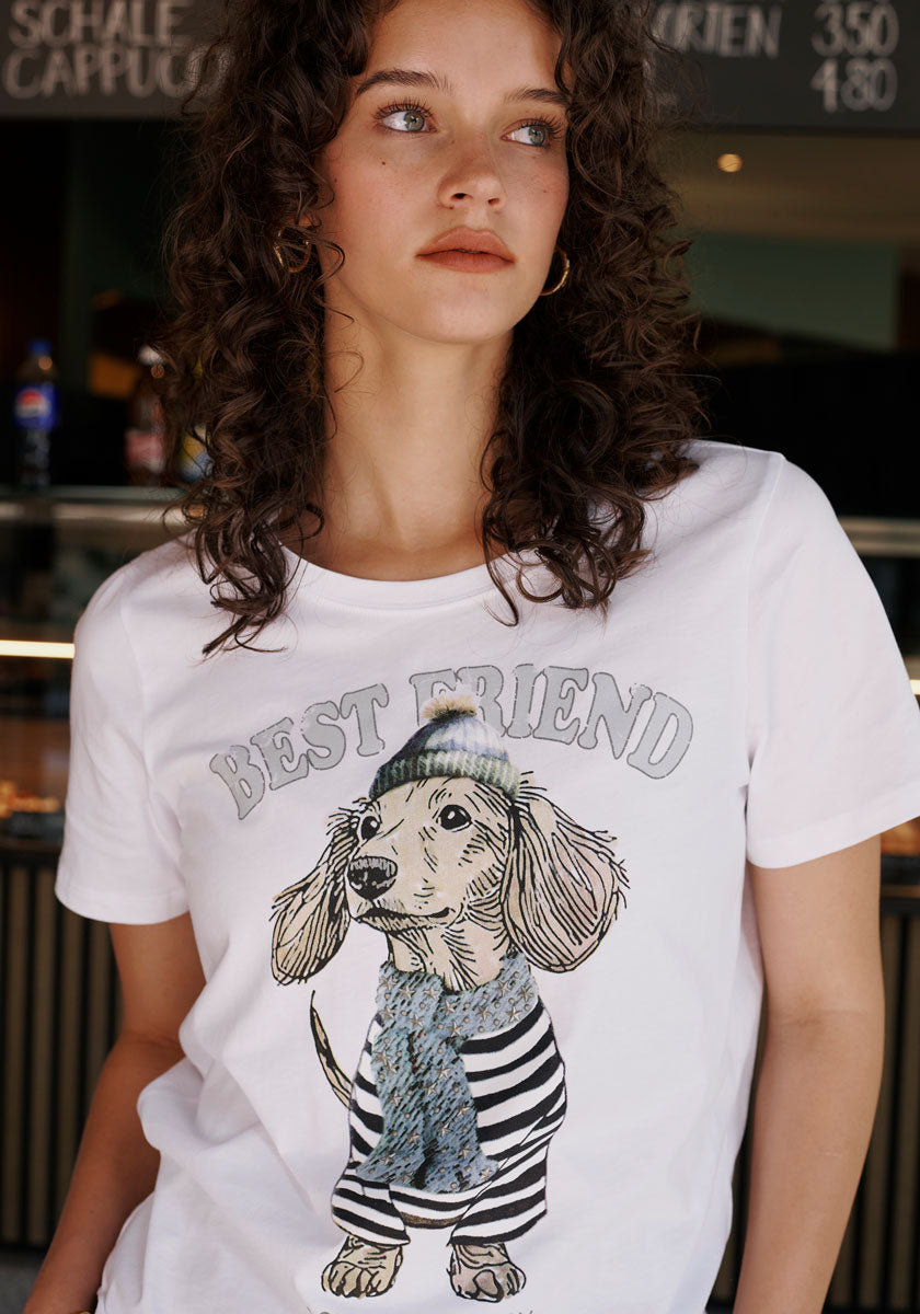 A woman with curly brown hair and hoop earrings wears a Princess Goes Hollywood white cotton T-shirt featuring an illustrated dachshund in a striped shirt and blue scarf, with "Best Friend" above. She stands indoors, a blurred menu board behind her.