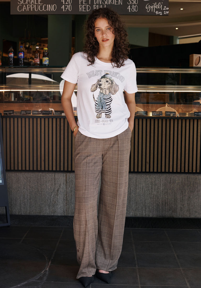 A woman with curly hair wears a Princess Goes Hollywood cotton T-shirt featuring a dachshund print and “Best Friend,” paired with loose brown plaid pants and black shoes, standing relaxed by a cafe counter with hands in her pockets.