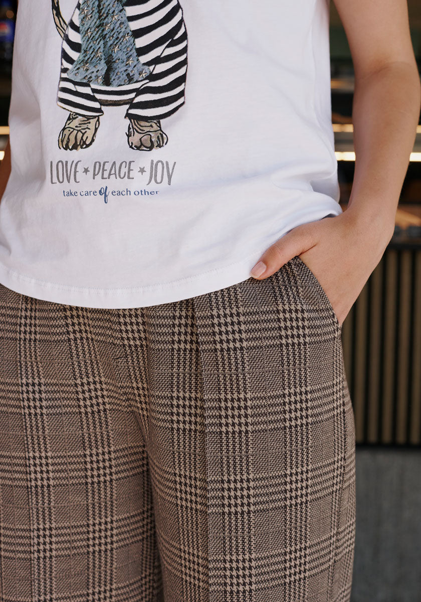 A person is seen from torso to upper legs wearing Princess Goes Hollywood’s wide-leg check trousers and a white T-shirt with an illustrated animal and “LOVE PEACE JOY, take care of each other”; the trousers have a comfy elastic waistband.
