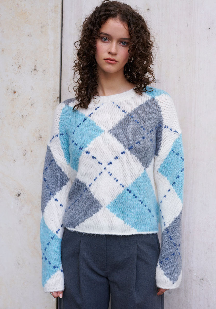 A young woman with curly brown hair models a Princess Goes Hollywood wool-alpaca blend sweater with a large light blue, gray, navy, and white argyle pattern, paired with high-waisted gray pants. She stands before a light textured wall, looking at the camera.