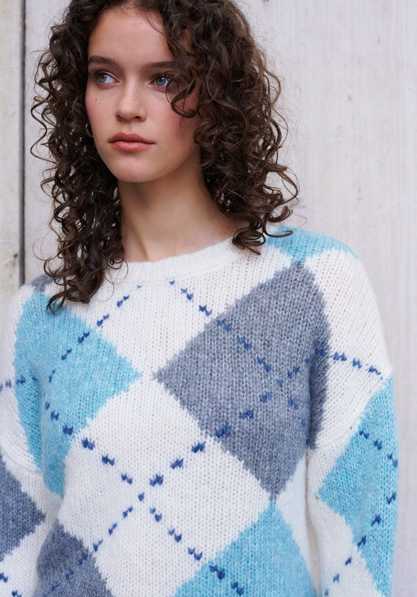 A young woman with curly brown hair and blue eyes wears a Princess Goes Hollywood Strickpullover with a bold blue-white argyle pattern in wool-alpaca blend, standing before a light wooden wall, gazing to the side with a neutral expression.