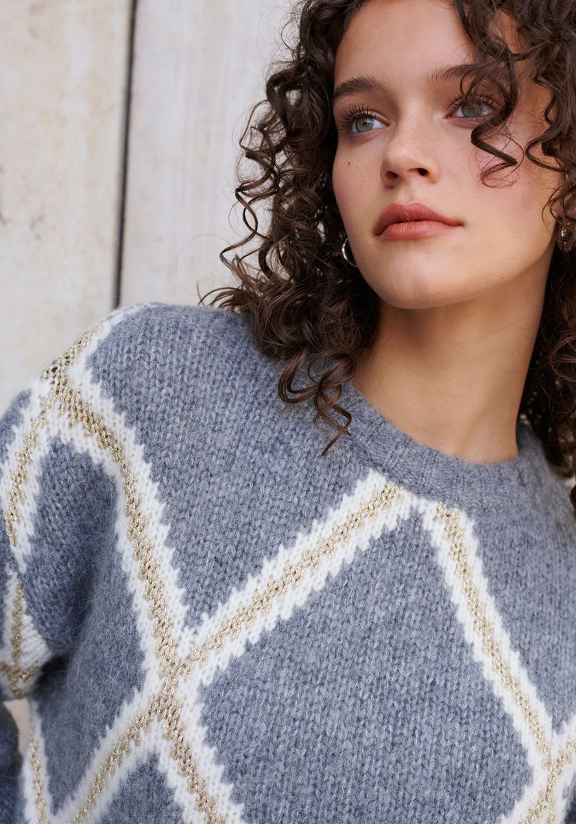 A young woman with curly brown hair and blue eyes gazes upward, wearing a soft gray Princess Goes Hollywood Alpaka-Mix Strickpullover with a white and beige argyle pattern. A light wood background enhances the cozy, wintery vibe.