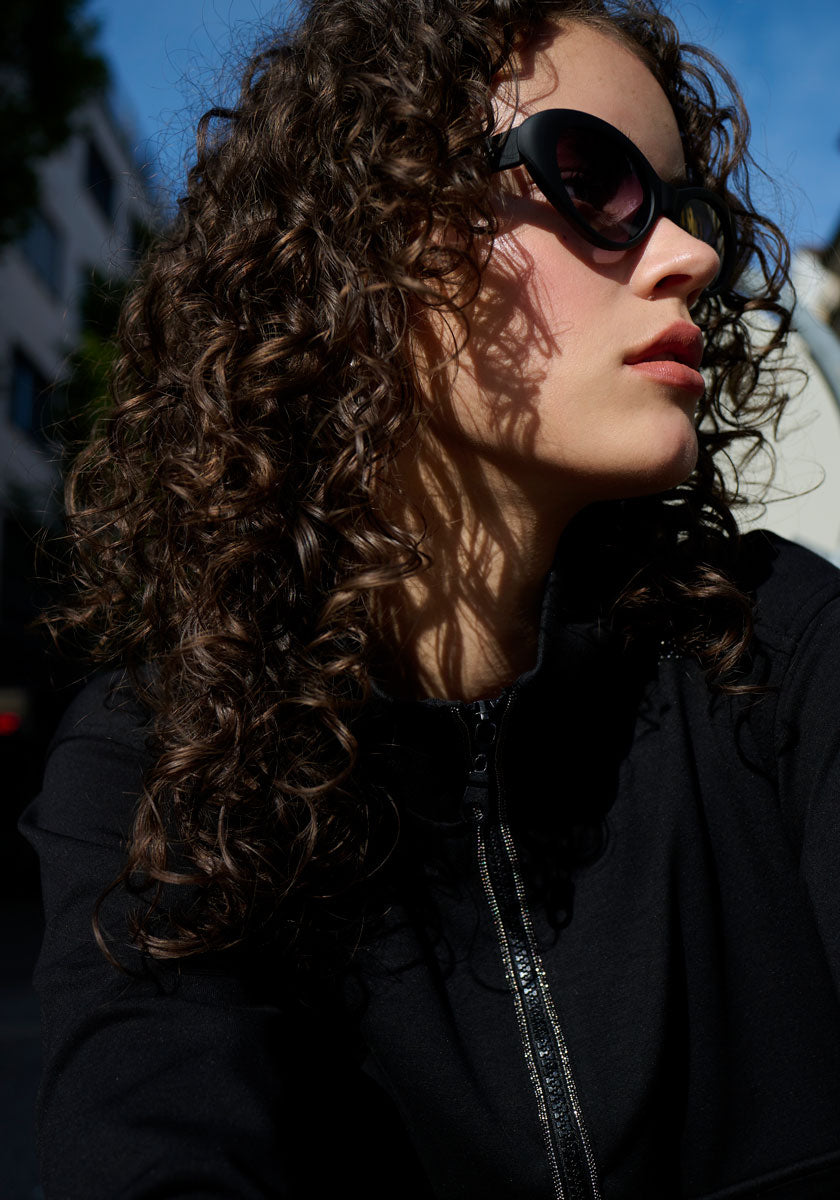 A woman with long, dark curls wears large black sunglasses and a black Princess Goes Hollywood Sweatjacke with subtle rhinestone details. She gazes to the side, sunlight accentuating her hair as the blurred cityscape and sky form the backdrop.