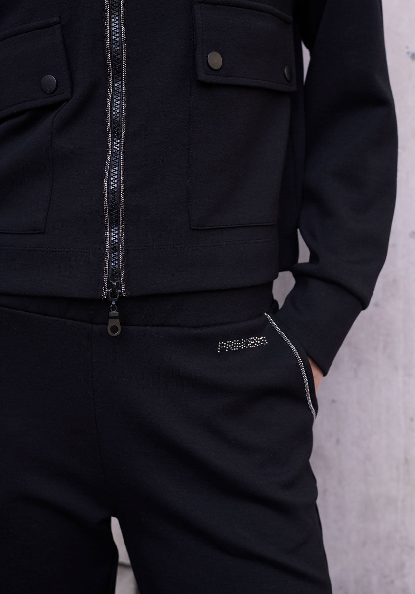 A person in Princess Goes Hollywood: a black Sweatjacke with front pockets and metallic snap buttons on the flaps, paired with matching black pants featuring a small Strassverzierung/logo on the left pocket.