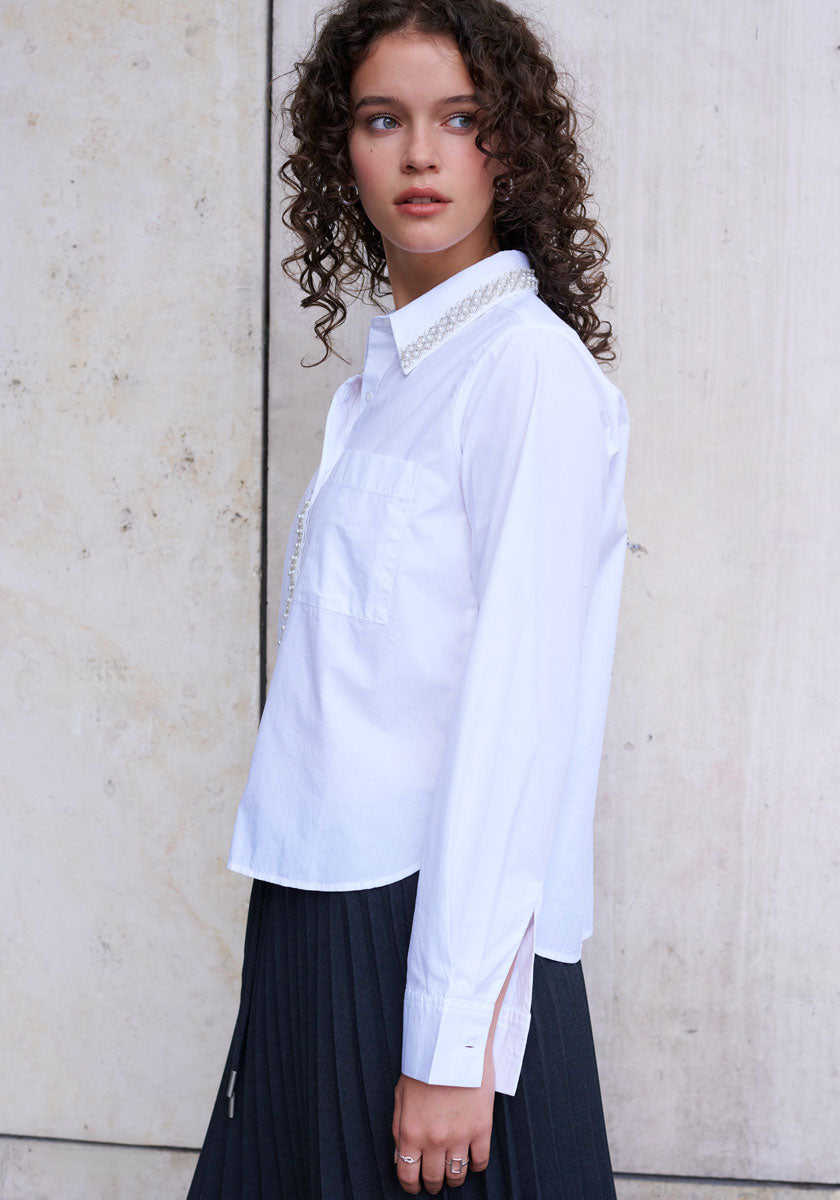 A young woman with curly brown hair wears a white button-up long-sleeve shirt and a black Plissee Rock mit elastischer Taille by Princess Goes Hollywood. She stands sideways, looking over her shoulder, against a textured light-colored wall.