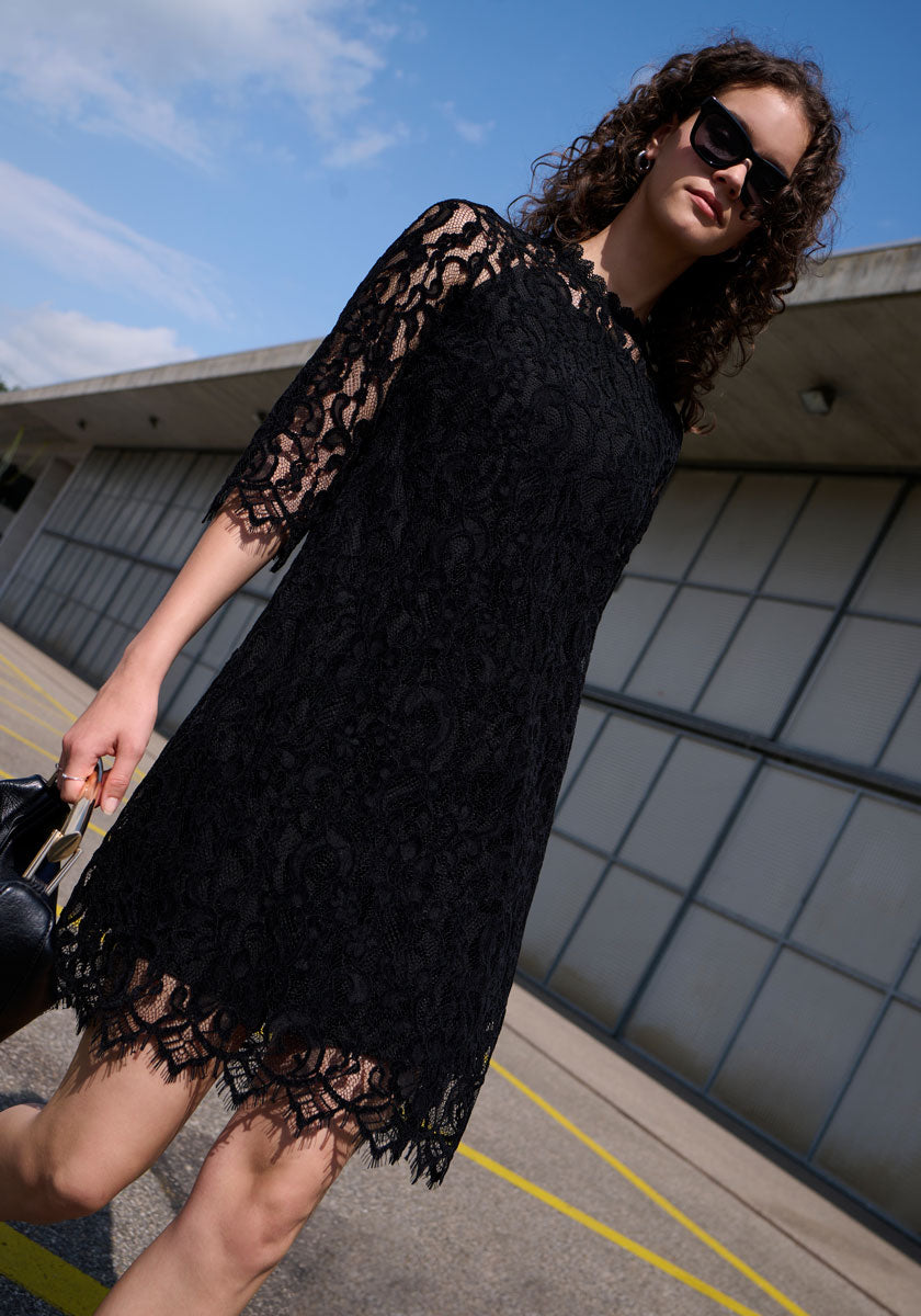 A woman with curly hair and sunglasses wears a black Princess Goes Hollywood Häkelspitze Kleid mit 3/4-Ärmel und Stehkragen, standing confidently outdoors with a black handbag. The sky is blue and a modern building is in the background.