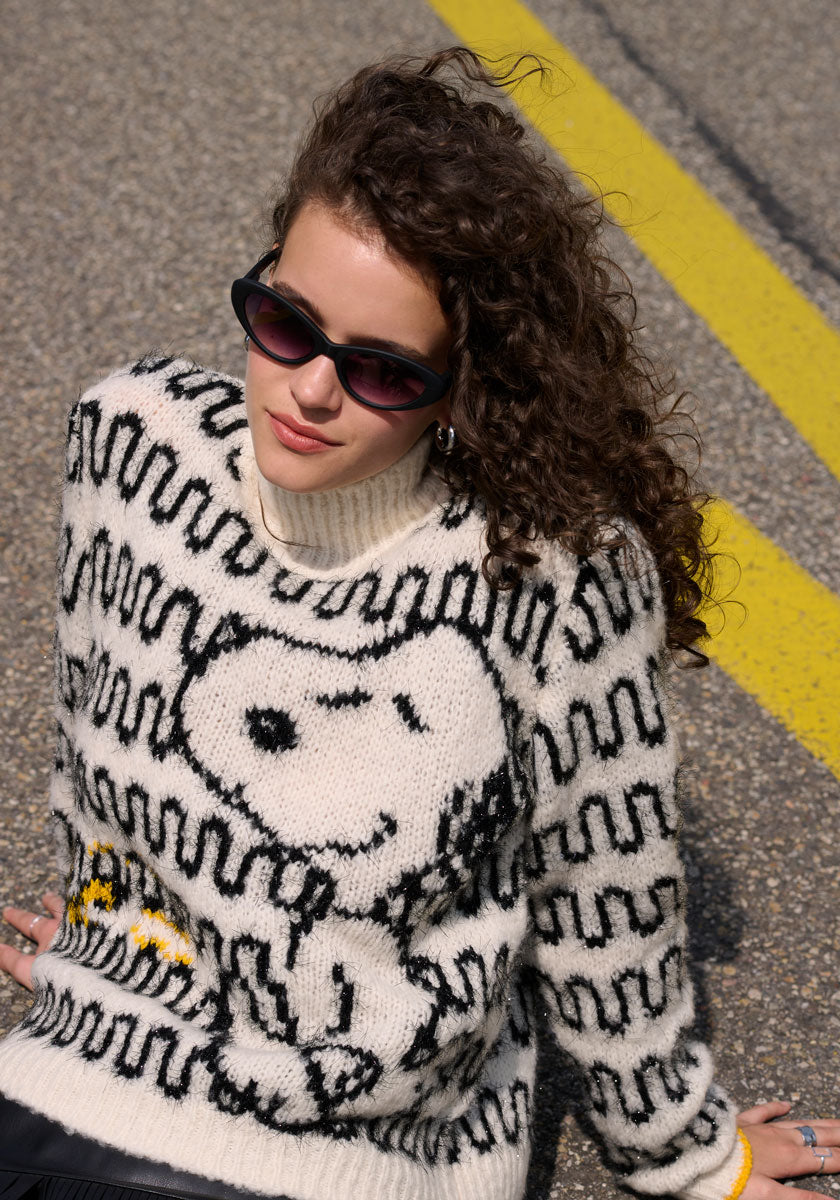 A woman in sunglasses sits on the ground, wearing a cozy Princess Goes Hollywood Wollmix Grobstrickpullover featuring a playful Snoopy-Motiv.
