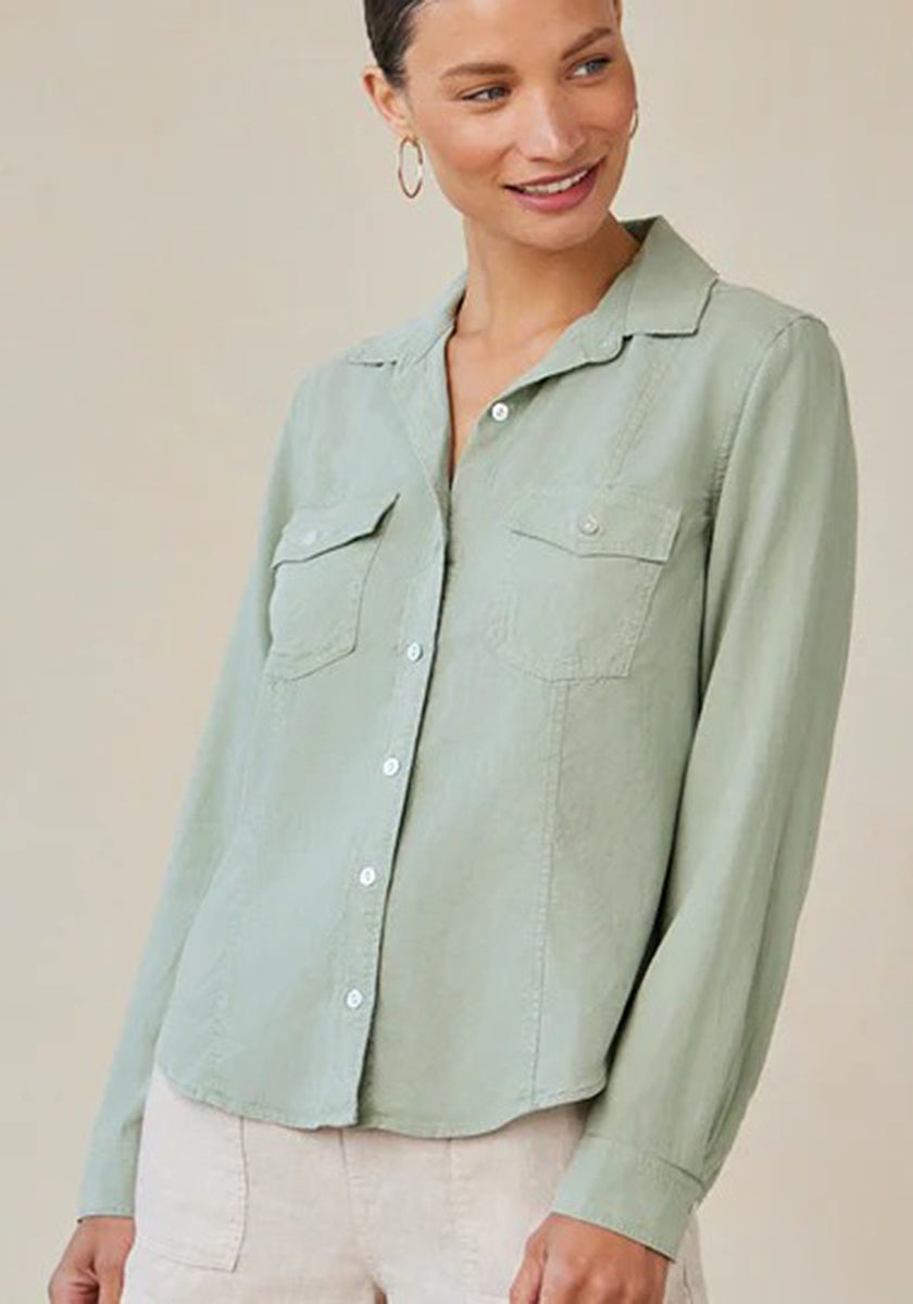 A woman wears a light sage green Bella Dahl Damen Hemdbluse with long sleeves and two chest pockets, paired with light-colored pants. She stands against a beige background, smiling slightly to her left, her hair pulled back and hoop earrings on.