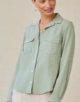 A woman wears a light sage green Bella Dahl Damen Hemdbluse with long sleeves and two chest pockets, paired with light-colored pants. She stands against a beige background, smiling slightly to her left, her hair pulled back and hoop earrings on.