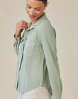 A woman with dark hair pulled back wears a light sage green Bella Dahl Lyocell Bluse im Jacken-Stil with chest pockets and long sleeves, looking down and smiling softly against a plain, neutral background.