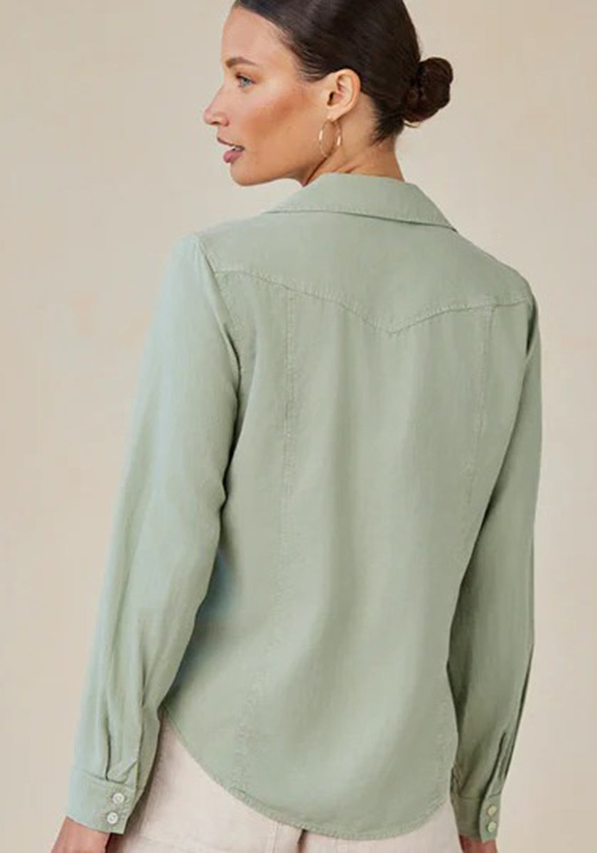 A woman with light skin and dark hair in a low bun is seen from the back, wearing a sage green Bella Dahl Lyocell Bluse im Jacken-Stil with a back yoke and light-colored pants. She has hoop earrings and stands against a neutral background.