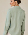 A woman with light skin and dark hair in a low bun is seen from the back, wearing a sage green Bella Dahl Lyocell Bluse im Jacken-Stil with a back yoke and light-colored pants. She has hoop earrings and stands against a neutral background.
