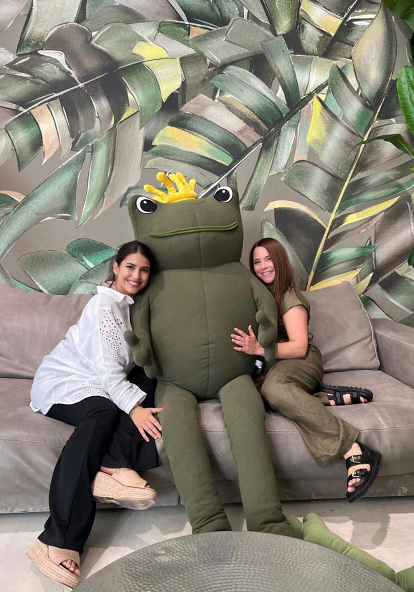 Two women sitting on a gray couch smile and hug a large green stuffed frog with a yellow crown. The background features a decorative wall with a leafy tropical pattern. Both women appear happy and relaxed, dressed in casual clothes and sandals.