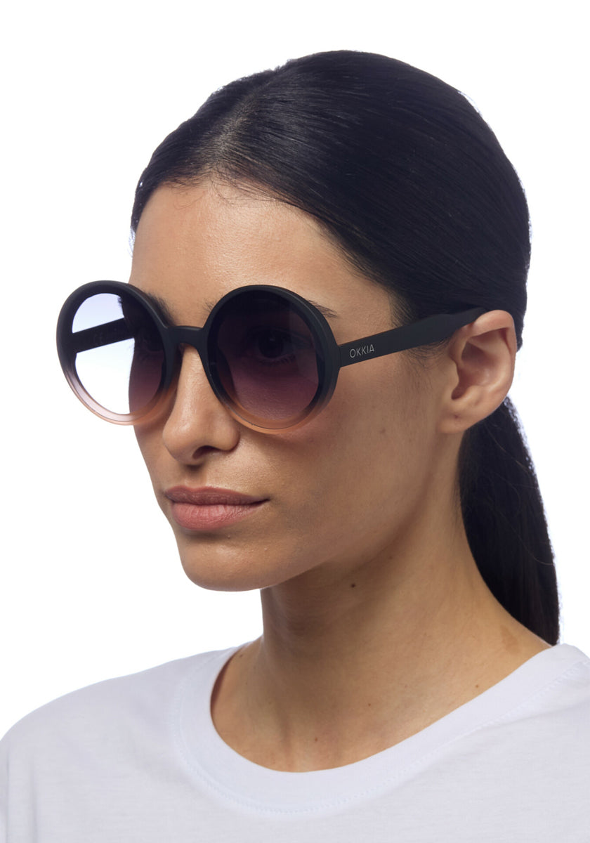 A woman with straight, dark hair tied back wears Okkia’s Oversize-Sonnenbrille mit UV 400-Schutz und großem, rundem Design. Sie trägt ein weißes T-Shirt; das Okkia-Logo ist am linken Bügel vor schlicht weißem Hintergrund sichtbar.