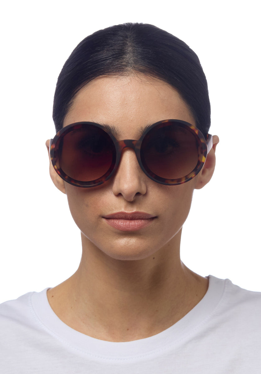 A woman with straight dark hair pulled back wears Okkias Oversize-Sonnenbrille with UV 400-Schutz and a white crew-neck shirt. She faces forward against a white background—ideal for stilbewusste Persönlichkeiten.