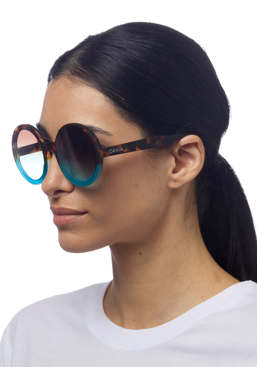 Eine Frau mit langen dunklen Haaren trägt Okkia Oversize-Sonnenbrille mit UV400-Schutz, runden blau verlaufenden Gläsern und Tortoiseshell-Gestell – ein stilvoller Look im weißen Shirt vor weißem Hintergrund.