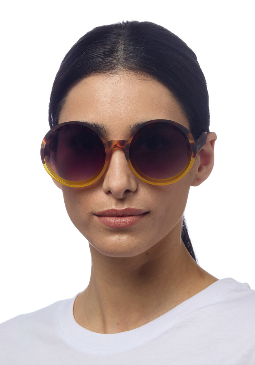 A woman with straight dark hair tied back wears a white shirt and Okkia Oversize-Sonnenbrille with brown-to-yellow faded frames and dark lenses—ideal for stilbewusste Persönlichkeiten—looking at the camera against a plain white background.