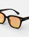 Nahaufnahme der schildpattfarbenen Sonnenbrille von Okkia mit orangefarbenen, verspiegelten Gläsern und quadratischem Design. Der dicke, rechteckige Rahmen ist mit einem weißen Kreuz, dem Okkia-Branding und UV-400-Schutz versehen und steht auf einem schlichten weißen Hintergrund.