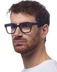 A man with short brown hair and a trimmed beard wears a white crew-neck shirt and black rectangular Okkia Bildschirmbrille for optimal comfort, facing left with a neutral expression against a plain white background.
