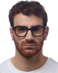 A man with short brown hair and a full reddish-brown beard wears large black Okkia Lesebrille in a soft-touch, square-frame design and a white crew-neck shirt, facing forward with a neutral expression against a plain white background.