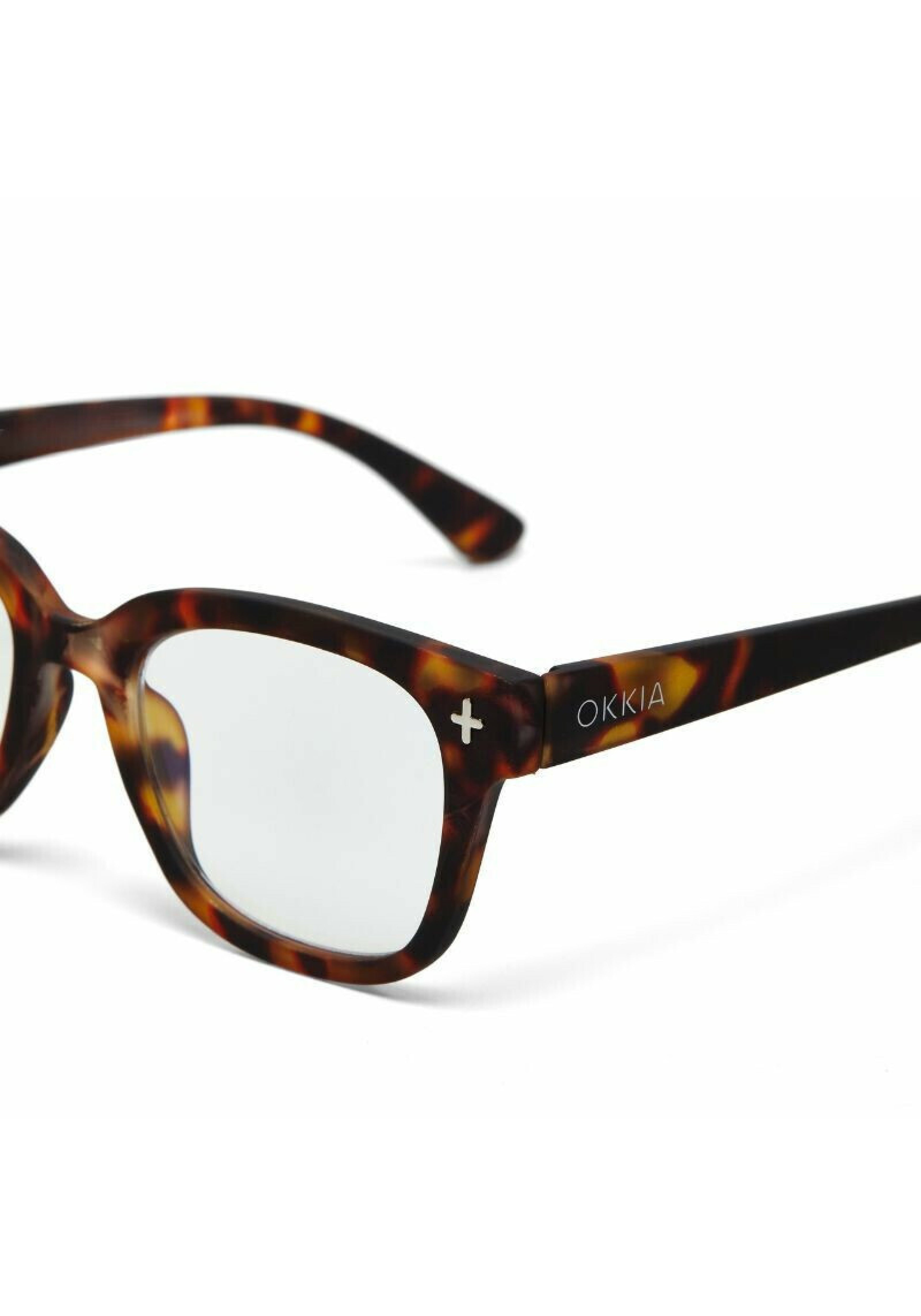 Close-up of tortoiseshell Okkia Blaulichtfilterbrille eyeglasses with thick, rounded frames. The left temple arm shows a white plus sign and “OKKIA.” Clear lenses provide screen comfort; background is plain white.