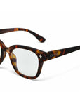 Close-up of tortoiseshell Okkia Blaulichtfilterbrille eyeglasses with thick, rounded frames. The left temple arm shows a white plus sign and “OKKIA.” Clear lenses provide screen comfort; background is plain white.
