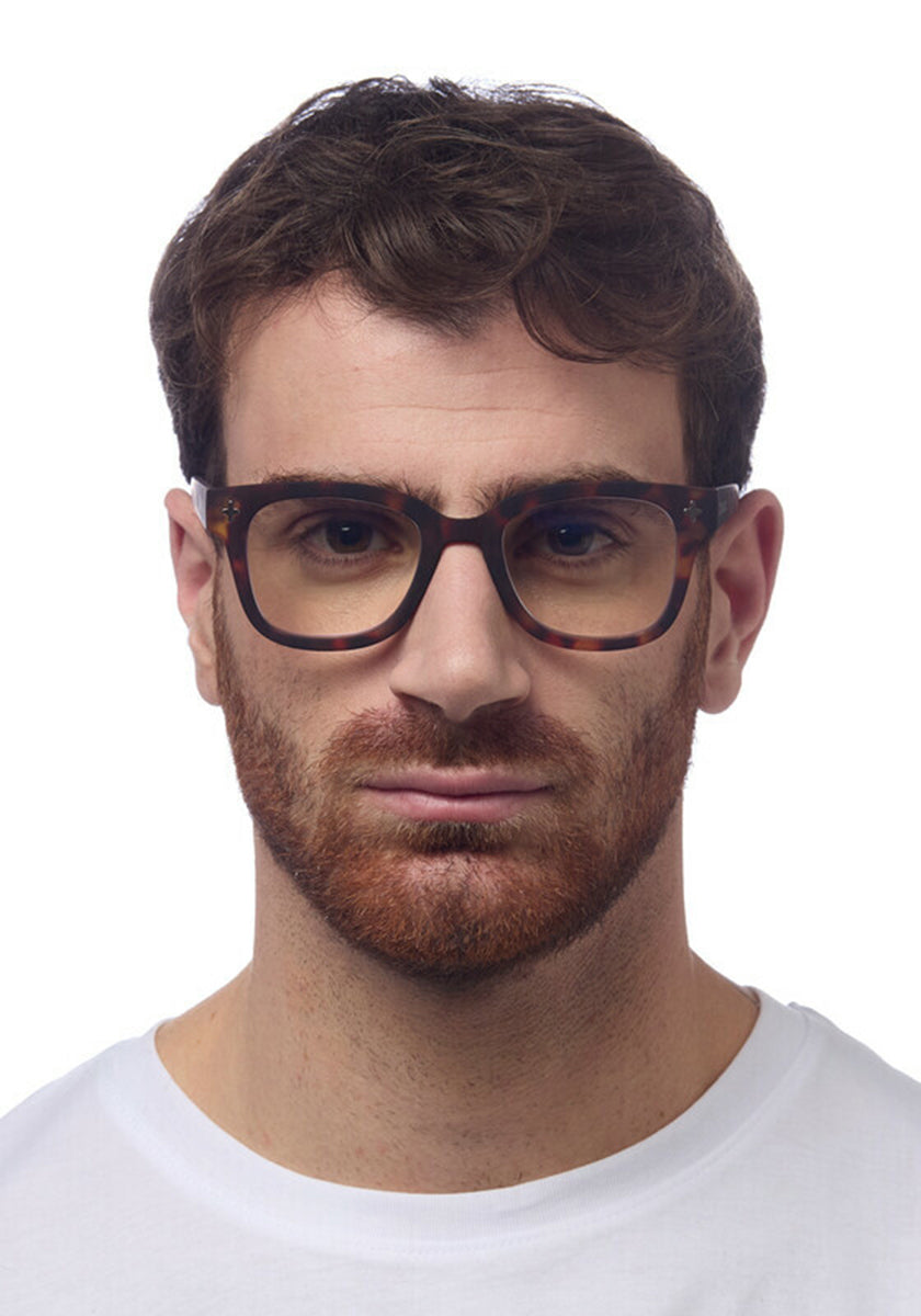 A man with short brown hair and a full reddish-brown beard wears Okkia dark-framed Bildschirmbrille for optimal Tragekomfort and a plain white crew-neck shirt, gazing ahead with a neutral expression against a white background.