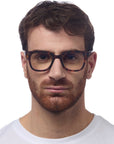 A man with short brown hair and a full reddish-brown beard wears Okkia dark-framed Bildschirmbrille for optimal Tragekomfort and a plain white crew-neck shirt, gazing ahead with a neutral expression against a white background.