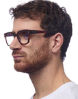 A man with short, wavy brown hair and a trimmed beard is shown in profile, wearing Okkia square-frame Lesebrille with a soft-touch frame. He pairs them with a plain white T-shirt against a white background.