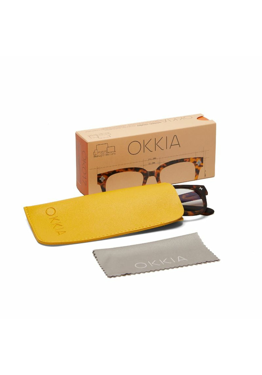 A pair of Okkia tortoiseshell square reading glasses partially in a yellow case, a gray cleaning cloth, and an Okkia branded box with product illustrations—all displayed on a white background.