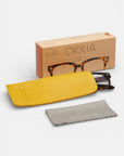Okkia-Schildpatt-Lesebrille mit quadratischem Rahmen, teilweise in einem gelben Etui, darunter ein graues Putztuch, ausgestellt vor einer beigen Okkia-Schachtel mit Brillengrafik. Alles vor einem schlichten weißen Hintergrund.