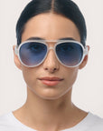Eine Person mit glattem, dunklem Haar trägt eine große Okkia Aviator-Sonnenbrille mit UV-400-Schutz, weißem Rahmen und blau getönten Gläsern. Sie hat einen neutralen Gesichtsausdruck und trägt ein schlichtes weißes Hemd vor einem hellen Hintergrund.