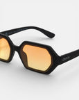 Nahaufnahme der schwarz gerahmten Okkia Andrea Sonnenbrille mit geometrischen, achteckigen, orange getönten Gläsern. Der dicke, matte Rahmen ist mit UV 400 Schutz und dem Okkia-Branding versehen und steht auf einem schlichten weißen Hintergrund.