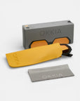 Eine Okkia-Sonnenbrille mit orangefarbenen Gläsern und geometrischem Design sitzt teilweise in einem gelben Etui vor einer grauen Okkia-Box. Ein graues OKKIA-Reinigungstuch liegt daneben, alles auf weißem Hintergrund.