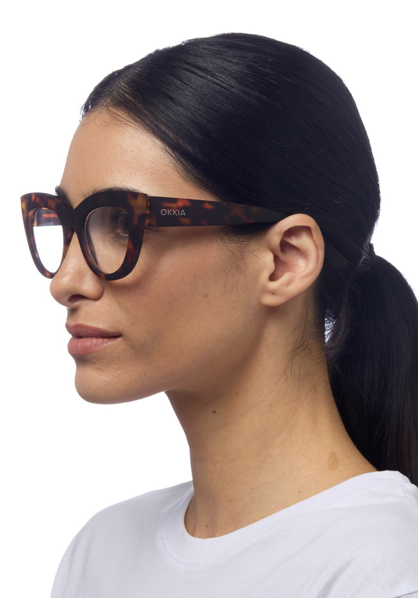 A woman with straight black hair in a low ponytail wears Okkia cat-eye reading glasses in Schildpattmuster, dressed in a white crew-neck top and facing left against a plain white background.