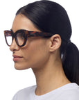 A woman with straight black hair in a low ponytail wears Okkia cat-eye reading glasses in Schildpattmuster, dressed in a white crew-neck top and facing left against a plain white background.