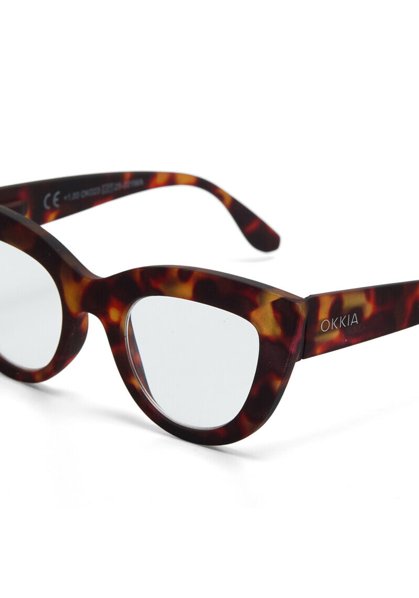 Close-up of Okkia eyeglasses with a bold cat-eye design in brown and amber tortoiseshell. Transparent lenses, with “OKKIA” in white on the left temple arm, set against a plain white background.