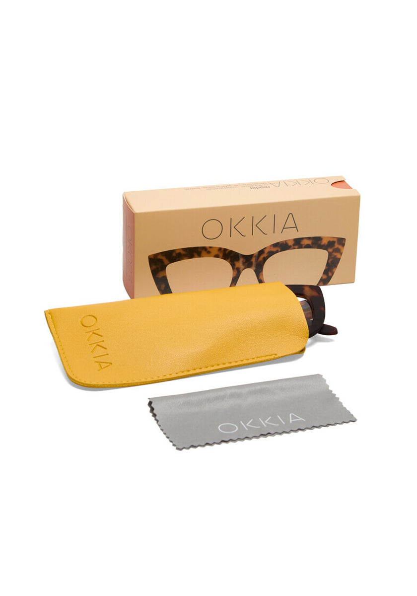 A pair of Okkia reading glasses in tortoiseshell, partially in a yellow case, with a grey OKKIA cleaning cloth and a branded cardboard box featuring an image of the glasses are displayed on a white background.