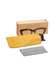 A pair of Okkia reading glasses in tortoiseshell, partially in a yellow case, with a grey OKKIA cleaning cloth and a branded cardboard box featuring an image of the glasses are displayed on a white background.