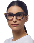 A woman with straight, dark hair tied back wears large, cat-eye Schildpattmuster Lesebrille from Okkia and a white shirt. She faces forward with a neutral expression against a plain white background.