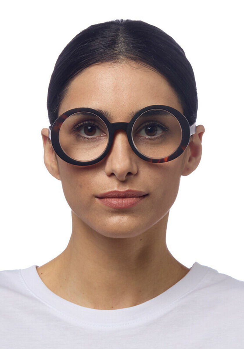 A woman with straight dark hair pulled back wears Okkia Oversize-Design round black Lesebrille eyeglasses. She has a neutral expression, is dressed in a plain white shirt, and the solid white background highlights her face and glasses.