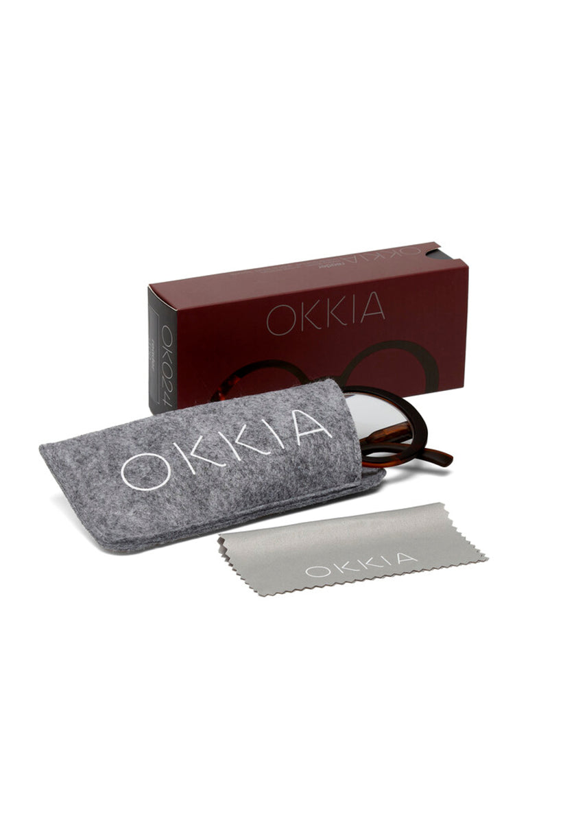 A pair of dark brown Okkia Oversize Lesebrille eyeglasses sits partly in a gray felt case with Okkia branding; a matching branded cloth lies in front, and behind them is a burgundy Okkia box, all on a white surface.