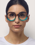 Eine Frau mit dunklem, nach hinten gezogenem Haar trägt Okkia Lesebrille im Oversize-Design mit rundem Schildpatt-Rahmen, der am unteren Ende in Teal übergeht. Sie hat einen neutralen Gesichtsausdruck, trägt ein schlichtes weißes Oberteil und steht vor einem hellen Hintergrund.