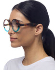 A woman with straight dark hair in a low ponytail wears large, round Okkia Lesebrille in an oversize design that fade to blue at the edges. She is shown in profile facing left, wearing a white T-shirt against a plain white background.