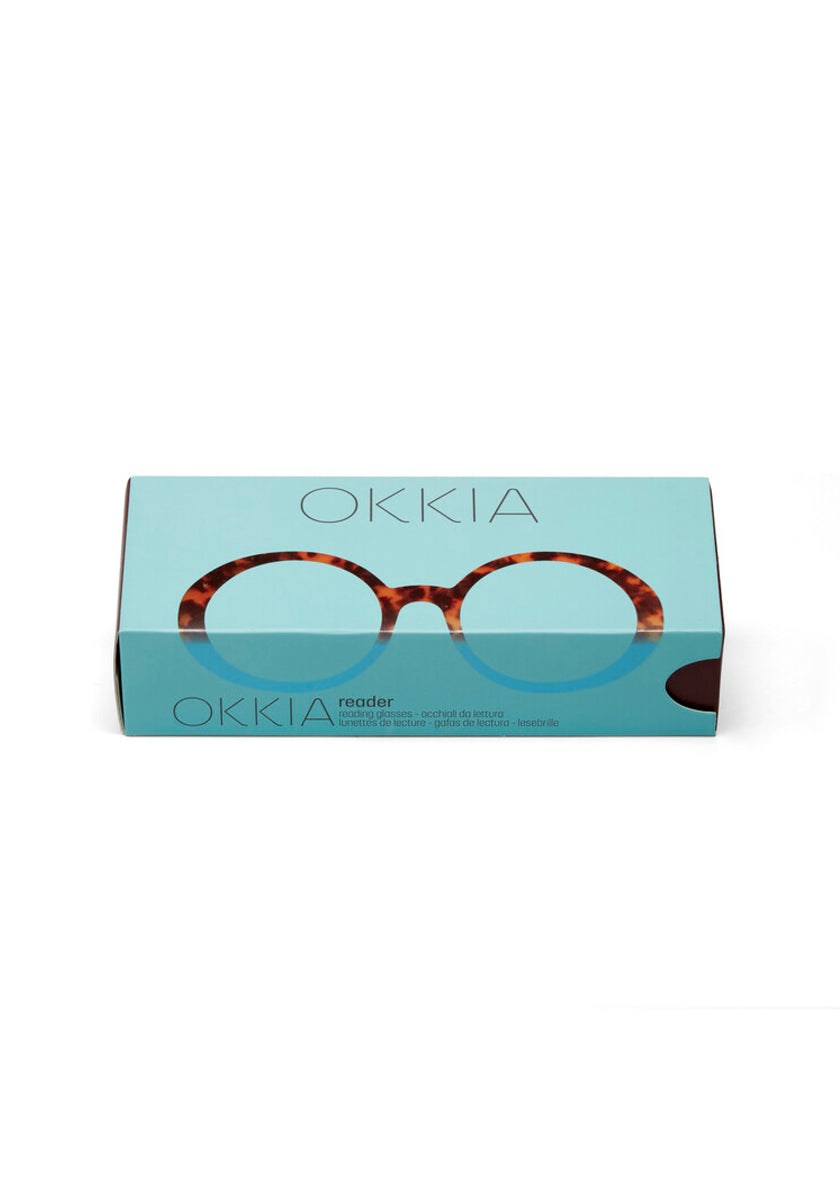 A rectangular turquoise Okkia box shows a photo of oversized tortoiseshell Lesebrille reading glasses on the lid. “Okkia” appears above and below the glasses, with product details in the lower left corner.
