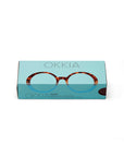 A rectangular turquoise Okkia box shows a photo of oversized tortoiseshell Lesebrille reading glasses on the lid. “Okkia” appears above and below the glasses, with product details in the lower left corner.