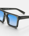 Diese rechteckige Sonnenbrille der Marke Okkia aus der VULCANO-Kollektion besticht durch einen glänzend schwarzen Rahmen und blaue Verlaufsgläser und verkörpert ein modernes Design mit übergroßen Proportionen. Das OKKIA-Logo ist in Weiß auf den linken Bügel aufgedruckt. Die Brille wird vor einem weißen Hintergrund präsentiert, wobei das Bild eng beschnitten ist, um die linke Seite und das Markenlogo hervorzuheben.
